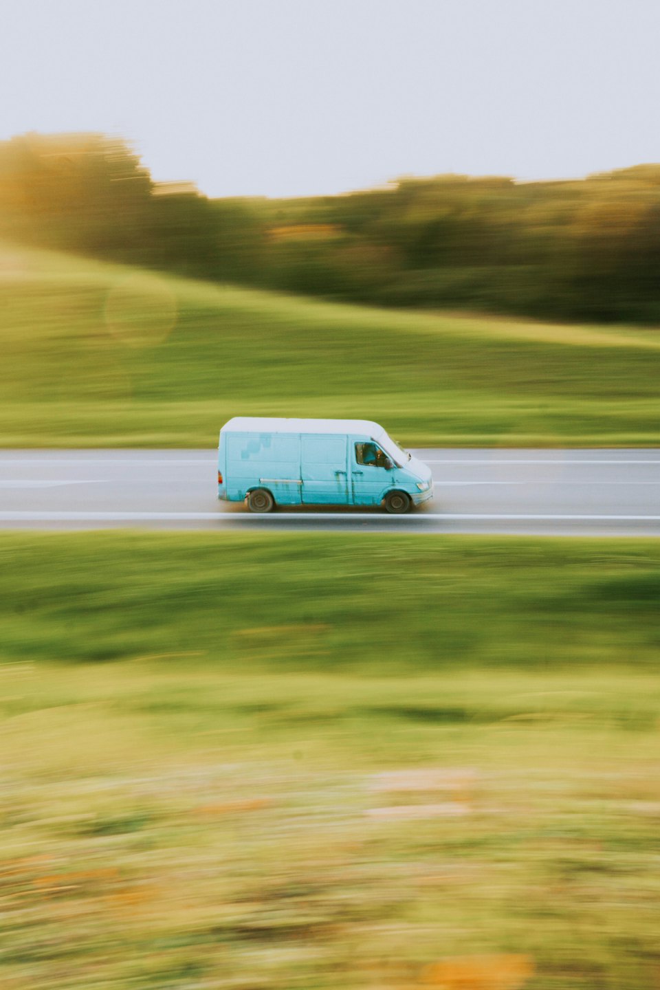 Light blue delivery van driving quickly along a rural road.